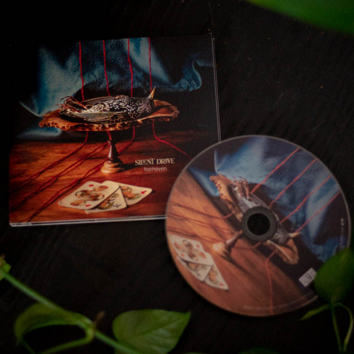 CD depicting a dead bird on a serving tray. Next to the tray is 3 playing cards. All of this is on top of a table.