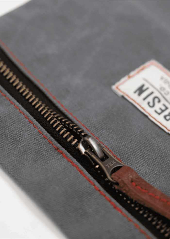 Gray fabric bag with a metal zipper and red stitching, featuring a visible label. The zipper pull is made of brown leather.