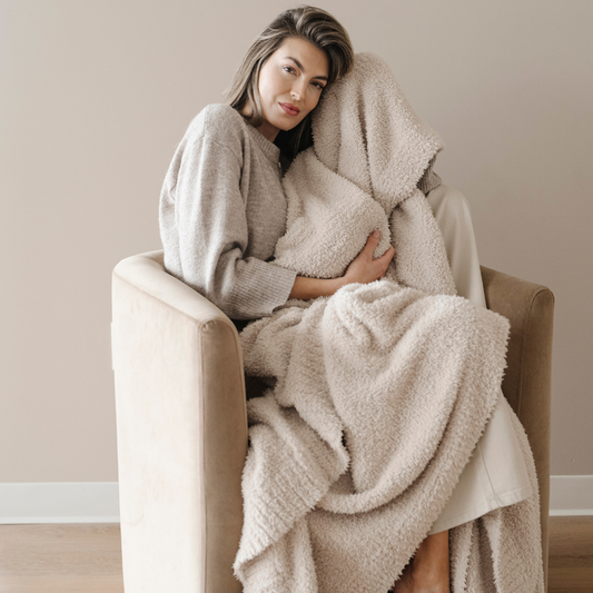 A woman is sitting in a chair and is hold a light tan Bamboni® blanket. This soft blanket is a luxury blanket and is a Saranoni blanket. This extra large blanket is super stretchy and breathable and is great for gifting, relaxing, or as a home decor staple. Enjoy this luxury blanket for years to come!