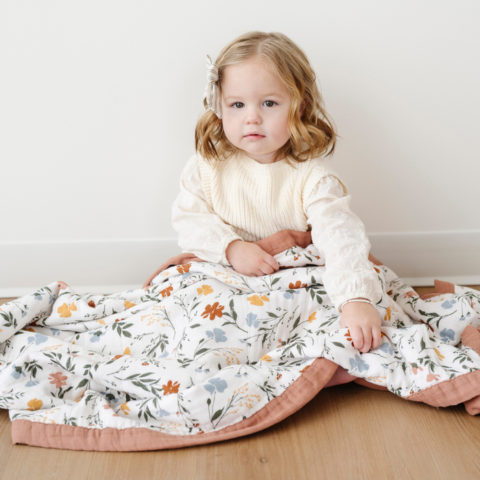 A little girl is sitting with a muslin quilt on her lap. This soft quilt is a Saranoni blanket that has a cute floral pattern on it. This quilt is a great little girl gift, baby gift, toddler gift, or addition to a nursery or bedroom. Enjoy this cozy blanket for years to come.