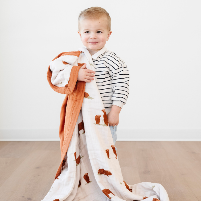 A little boy is holding a quilt with a highland cow pattern on it. This quilt is a Saranoni blanket and is a soft blanket. This quilt is a muslin fabric, which makes it a great baby gift, gift for toddlers, or addition to a nursery or bedroom. Enjoy this soft quilt for years to coe.