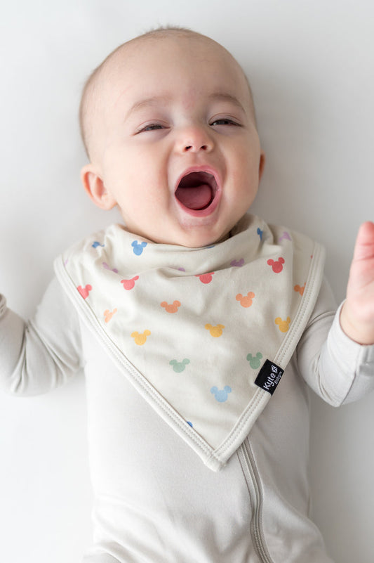 Baby in a cream outfit wearing a printed bib with Mickey Mouse heads in pastel colors