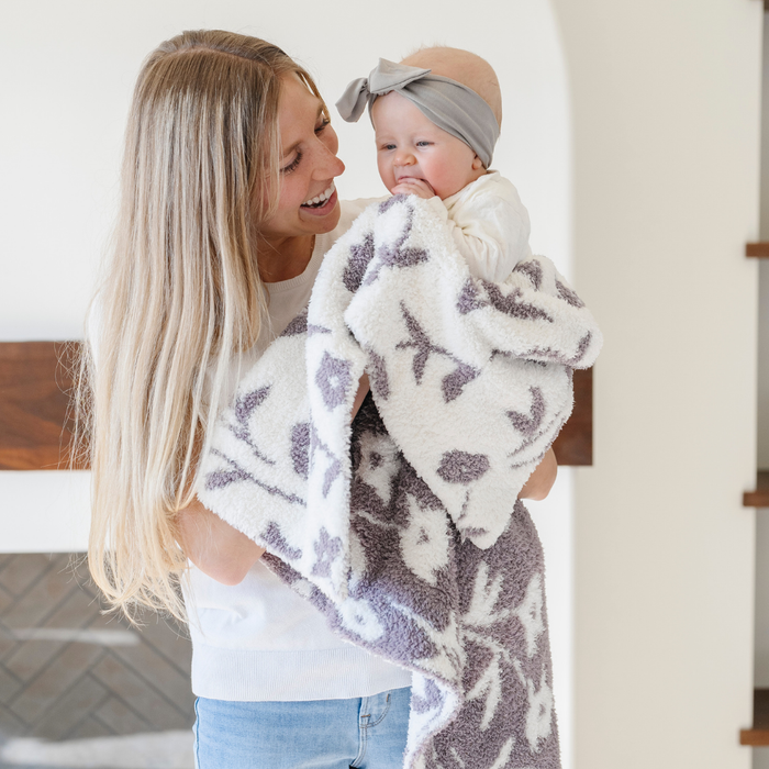 A mother smiles and holds her happy baby girl who is holding a double-layer Bamboni® baby blanket. The plush, breathable blanket features a two-tone purple and white floral design, offering ultimate softness and warmth. Made from Saranoni’s signature stretchy, cloud-like Bamboni® fabric, this luxury baby blanket provides comfort for naps, tummy time, and on-the-go snuggles. The soft blanket is a receiving blanket and Saranoni blanket.