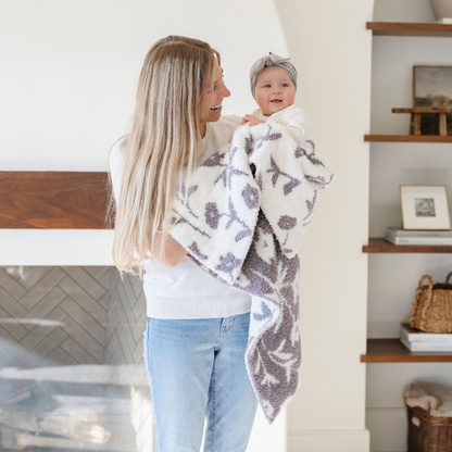 A mother smiles and holds her baby girl and a double-layer Bamboni® baby blanket. The plush, breathable blanket features a two-tone purple and white floral design, offering ultimate softness and warmth. Made from Saranoni’s signature stretchy, cloud-like Bamboni® fabric, this luxury baby blanket provides comfort for naps, tummy time, and on-the-go snuggles. The soft blanket is a receiving blanket and Saranoni blanket.