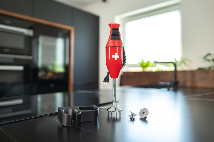 Der Stabmixer Unold ESGE-Zauberstab® E 120 Select Schweiz Rot mit Schweizer Kreuz steht auf einer schwarzen Küchentheke mit mehreren Aufsätzen und einer sonnendurchfluteten modernen Küche im Hintergrund.