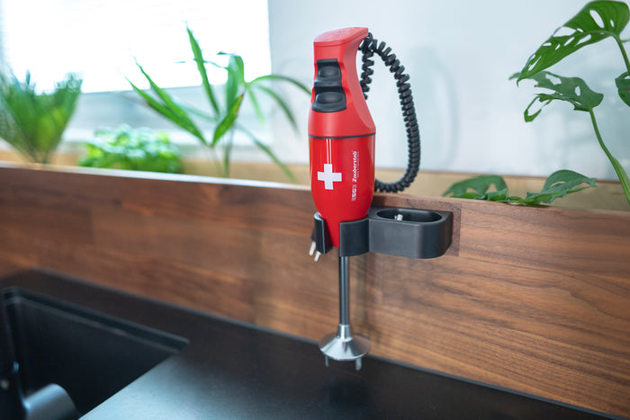 Der Unold ESGE-Zauberstab® E 120 Select Schweiz Rot, ein roter Stabmixer mit weißem Kreuz, ist an einer hölzernen Küchenwand in der Nähe von Grünpflanzen und einer schwarzen Arbeitsplatte montiert.