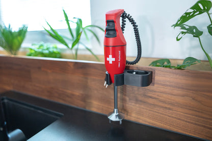 Der Unold ESGE-Zauberstab® E 120 Select Schweiz Rot, ein roter Stabmixer mit weißem Kreuz, ist an einer hölzernen Küchenwand in der Nähe von Grünpflanzen und einer schwarzen Arbeitsplatte montiert.