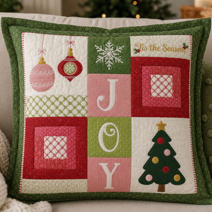 A square quilted throw pillow with a festive Christmas patchwork design, featuring the word 'JOY' in large letters, ornaments, a snowflake, a Christmas tree, and 'Tis the Season' text, resting on a beige sofa.