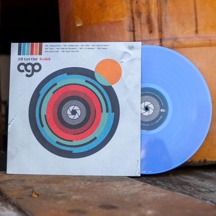 We see a vinyl cover that shows artwork that makes the cover look like a camera. In the middle is a lens and there are other color flares to give the camera style album art personality. Sticking out of the vinyl cover is a vinyl colored light blue. The background of this image is a rural setting showing the vinyl leaning against a barrel of sorts.