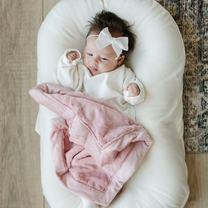 A Little Baby Girl with a white bow and white outfit and a Mini Saranoni Lush Blanket that is light pink. The specific color is called Ballet Slipper.