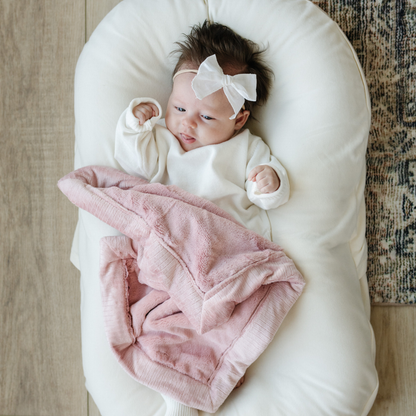 A Little Baby Girl with a white bow and white outfit and a Mini Saranoni Lush Blanket that is light pink. The specific color is called Ballet Slipper.