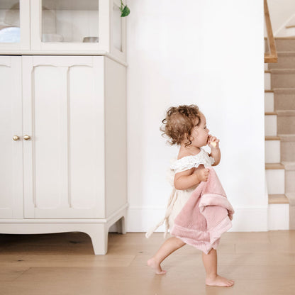A little girl wearing a white outfit walking through a house holding a Mini Saranoni Lush Blanket that is light pink. The specific color is called Ballet Slipper.