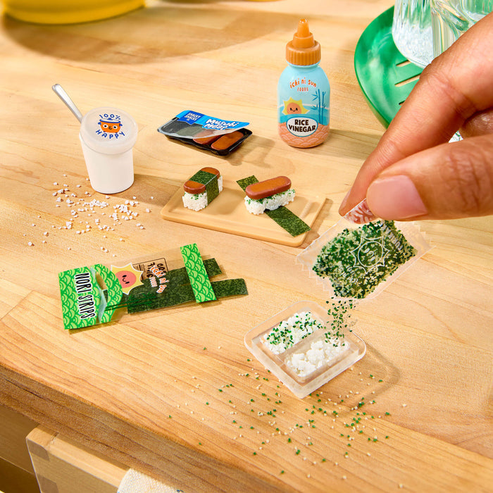 A close-up of a hand placing miniature  food items on a light wood surface, including small sushi pieces, a cup with a spoon, a bottle labeled 'Rice Vinegar,' and packaged snack pieces.
