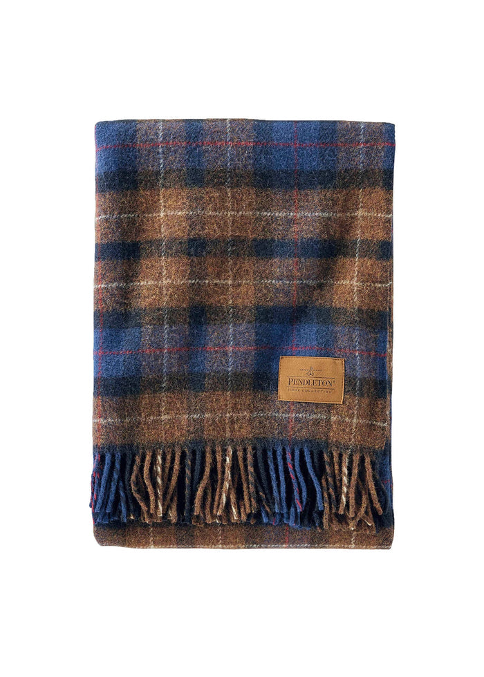 A folded Pendleton wool blanket with a blue and brown plaid pattern and fringe, featuring a leather logo patch on the front.