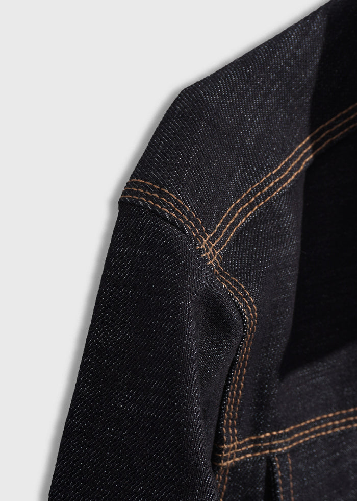 Close-up of black denim fabric with orange stitching, highlighting texture and seam details on a light gray background.