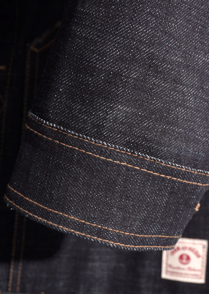 Close-up of a denim jacket sleeve with visible stitching and a small label at the bottom, highlighting texture and craftsmanship.