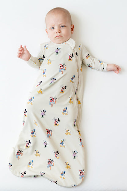 Infant laying on a light neutral surface wearing the Sleep Bag in Mickey Mouse and Friends 0.5 with matching romper