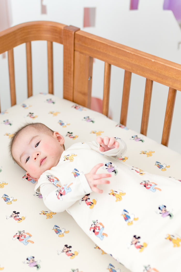 Baby in a wooden crib with a Mickey Mouse and Friends pattern wearing a matching sleep sack