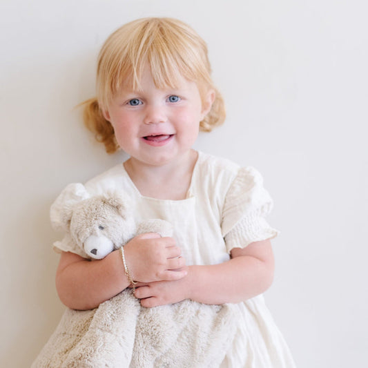 Adorable toddler girl in a white dress cuddling a Saranoni bear stuffed animal lovey. This ultra-soft plush lovey blanket features a cozy bear head and luxuriously soft fabric, designed to provide comfort, security, and soothing snuggles for babies and toddlers. Made with high-quality materials, the Saranoni lovey is perfect for newborns, infants, and young children as a bedtime companion or travel comfort item. Ideal baby shower gift or nursery essential, offering warmth and softness in a neutr