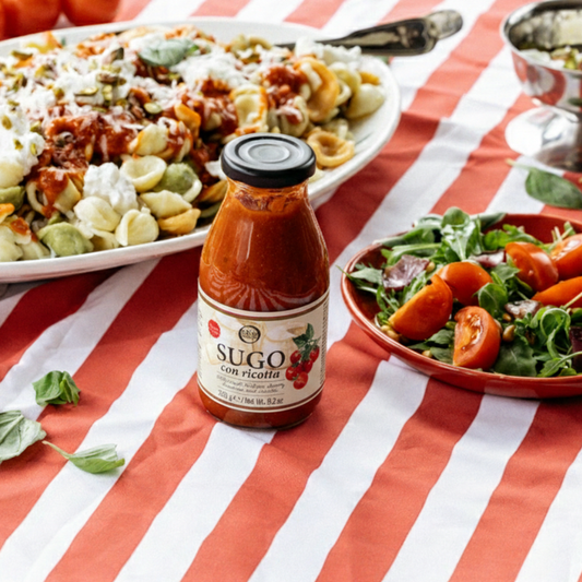 Bottle of Sugo con ricotta sauce on a red and white striped tablecloth with pasta and salad in the background.