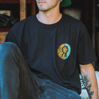 Wearing a black Hail The Sun Comfort Colors Moon T-Shirt with a yellow and blue circular graphic, a person with a forearm tattoo and backward green cap sits casually with one leg bent, capturing relaxed Hail The Sun style.
