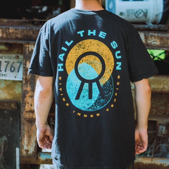 A person faces away from the camera in a black Hail The Sun Comfort Colors T-shirt with a vibrant sun circle graphic and DIVINE INNER TENSION text, standing before a rusted industrial background.