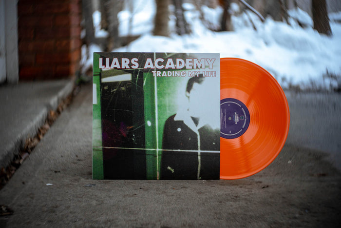 A bright orange vinyl by Liars Academy leans against its cover, which features Trading My Life text and a blurry black-green figure—inviting you to rediscover this classic from Liars Academy.