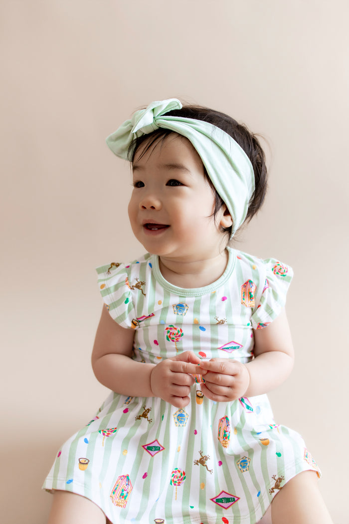 Child in a green and white striped twirl bodysuit dress with colorful Harry Potter patterns and a green headband on a beige background