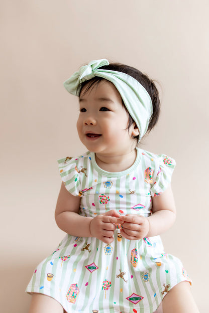 Child in a green and white striped twirl bodysuit dress with colorful Harry Potter patterns and a green headband on a beige background